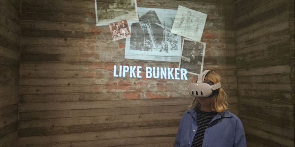A young lady wears a white VR headset, with the Lipke Bunker text and some old photos and papers projected on the wooden wall behind her.
