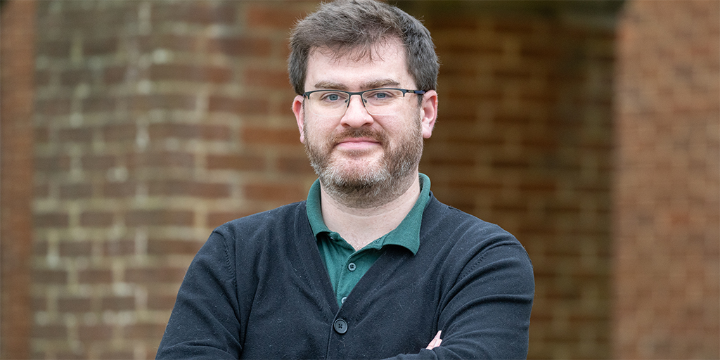 The photo shows an individual with short dark hair, glasses, and a beard. They are wearing a dark cardigan over a green collared shirt and have a confident, approachable expression. The background features a brick wall, adding a warm, rustic feel to the setting. Their arms are crossed, giving a relaxed yet professional posture.