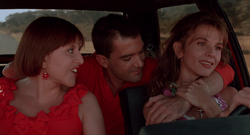 A woman drives a car. She is smiling Another woman sits next to her, is looking at her and smiling. A man leans forward from the backseat where he is sitting to embrace the woman who is driving. He is also smiling